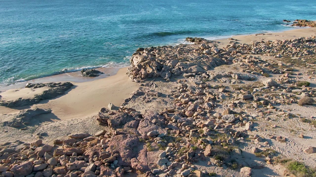 Aerial view over Buzzards Beach East Cape BCS Mexico with turquoise ocean, pale sand, rocky outcrops, and no visible people at the start of the clip.