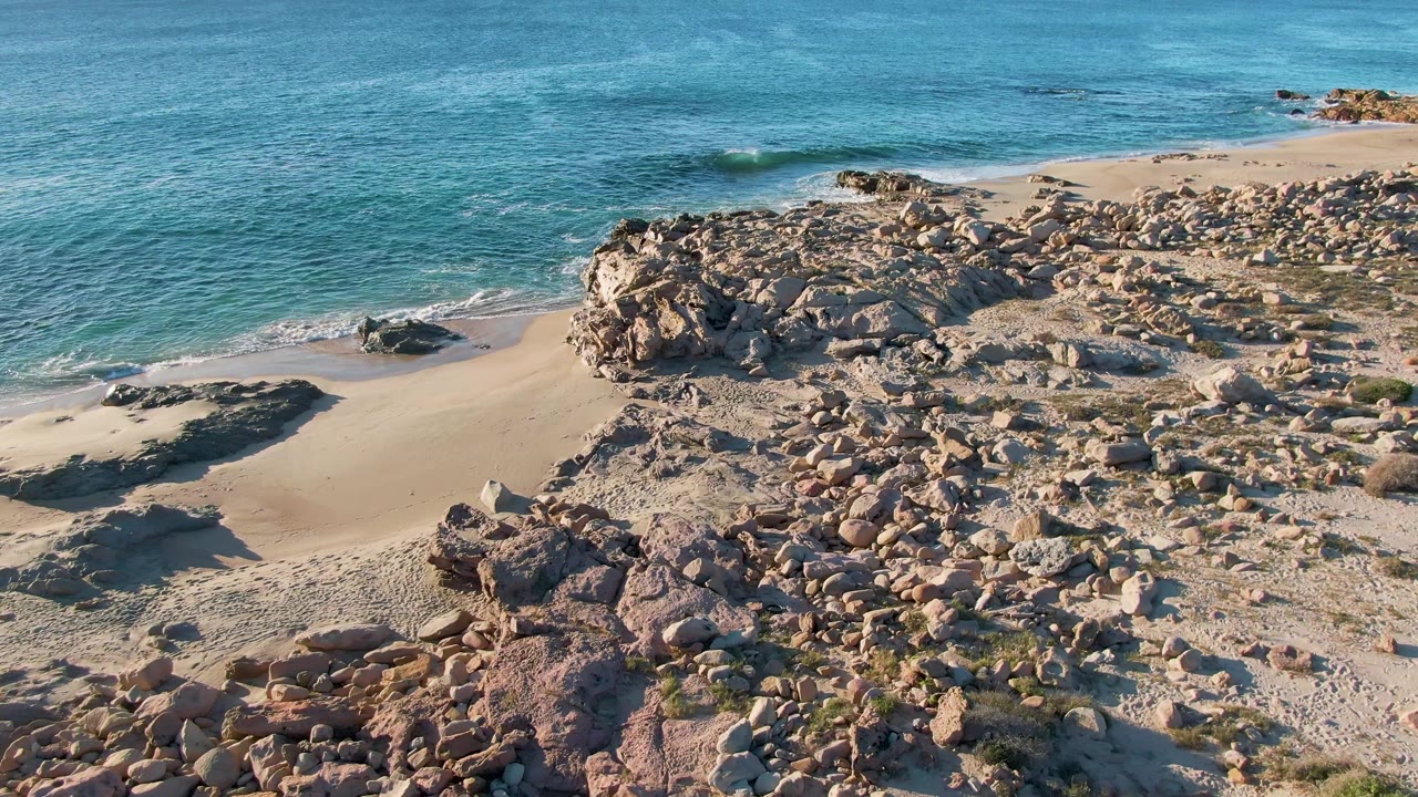 Drone view at Buzzards Beach East Cape BCS Mexico as small waves approach the sandy beach beside a broad cluster of tan rocks, with no visible people.