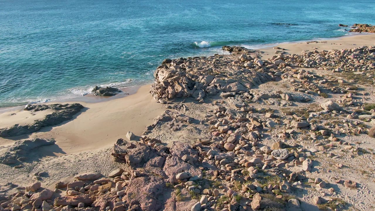 Aerial frame over Buzzards Beach East Cape BCS Mexico showing the rocky point filling the right side, blue ocean above it, and small white waves near the shore, with no visible people.