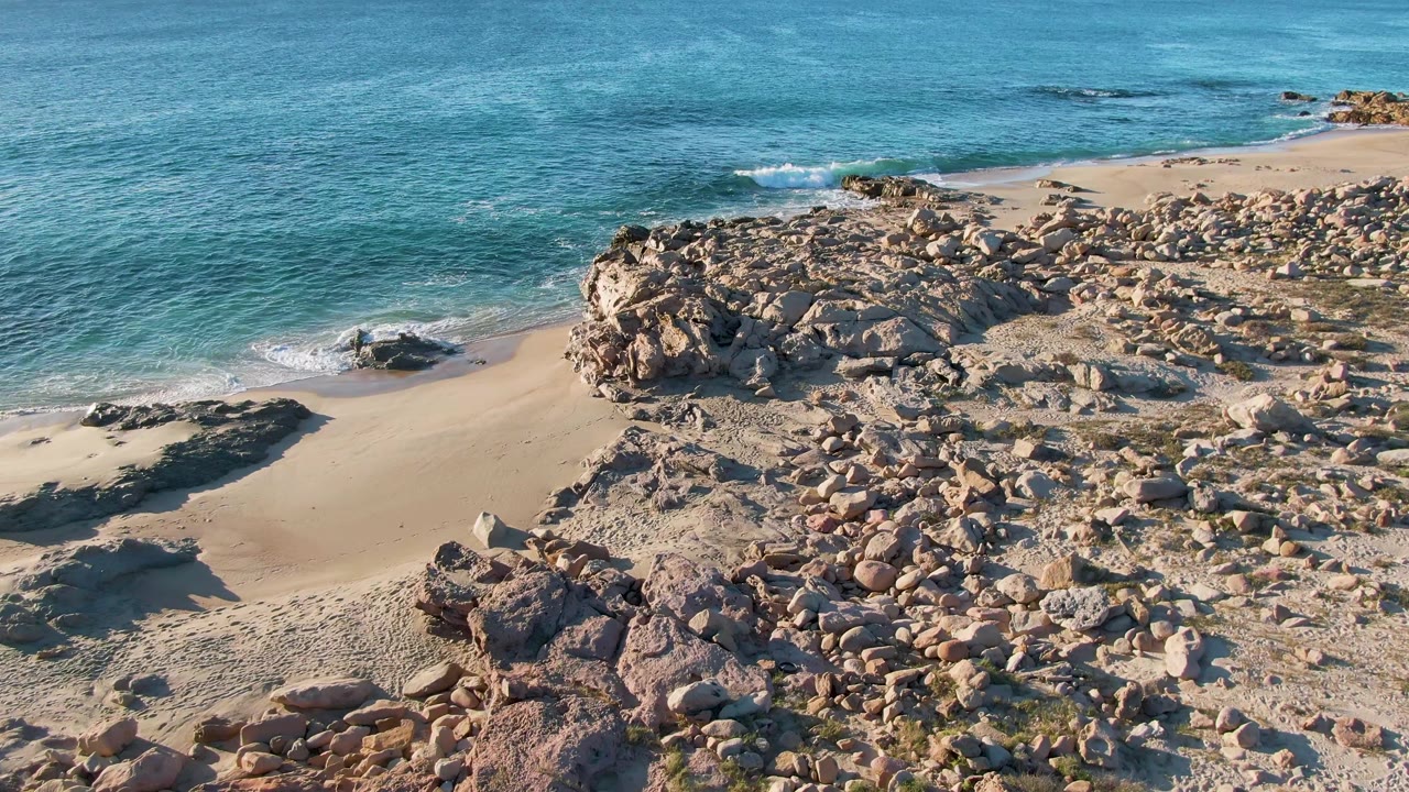 Aerial view of Buzzards Beach East Cape BCS Mexico with turquoise water breaking closer to the sandy cove, rocks spanning the foreground, and no visible people.