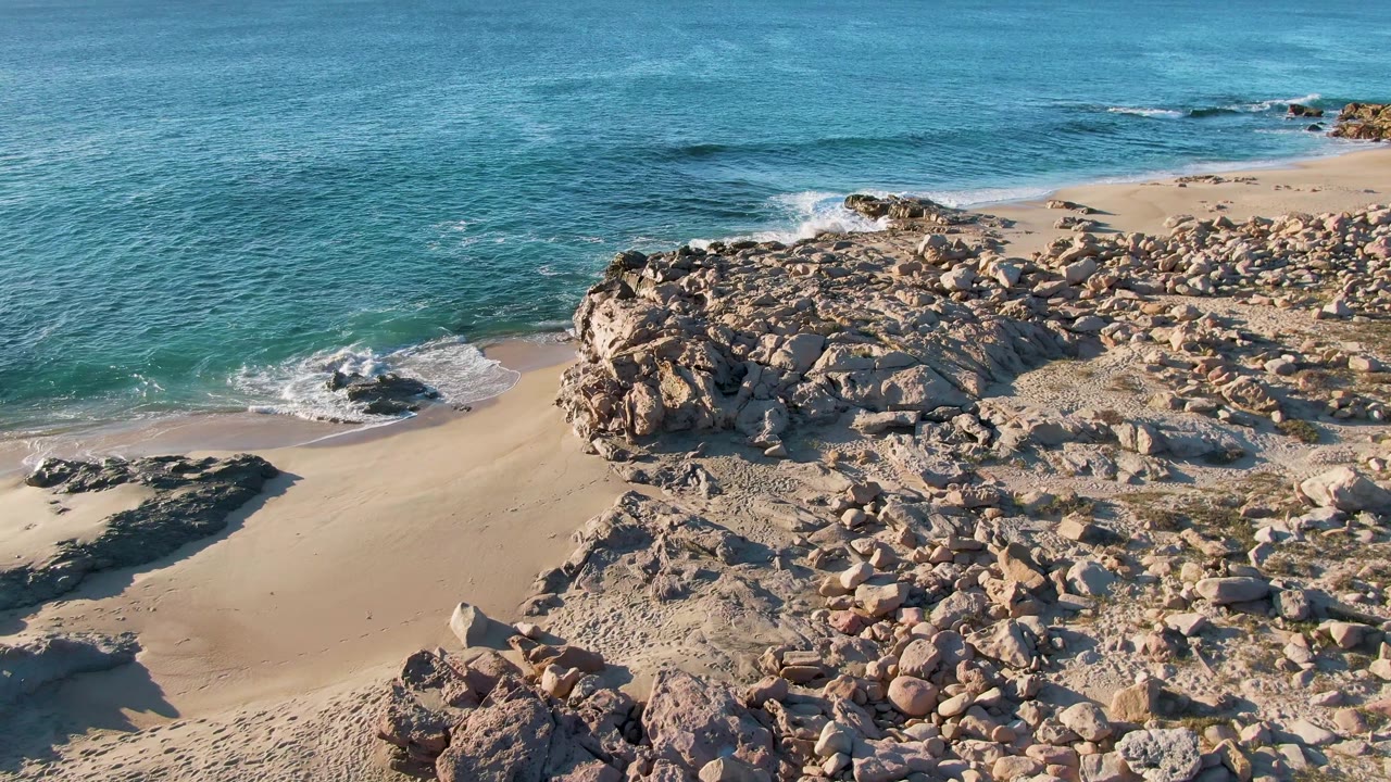 At 0:01, Buzzards Beach shoreline fills the frame as small white waves curl around dark rocks on sand with no visible people.