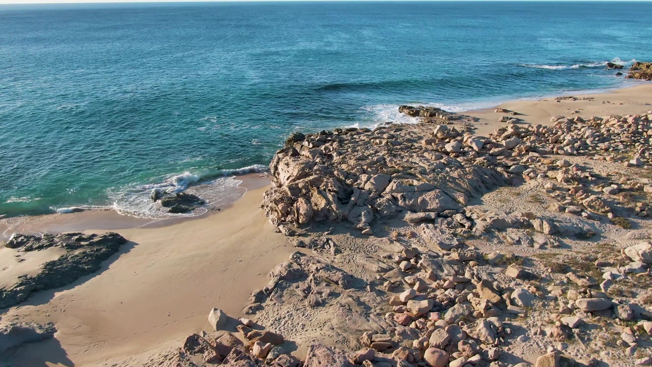 At 0:02, the aerial composition centers the rocky point and sandy cove at Buzzards Beach East Cape BCS Mexico, with blue ocean behind and no visible people.