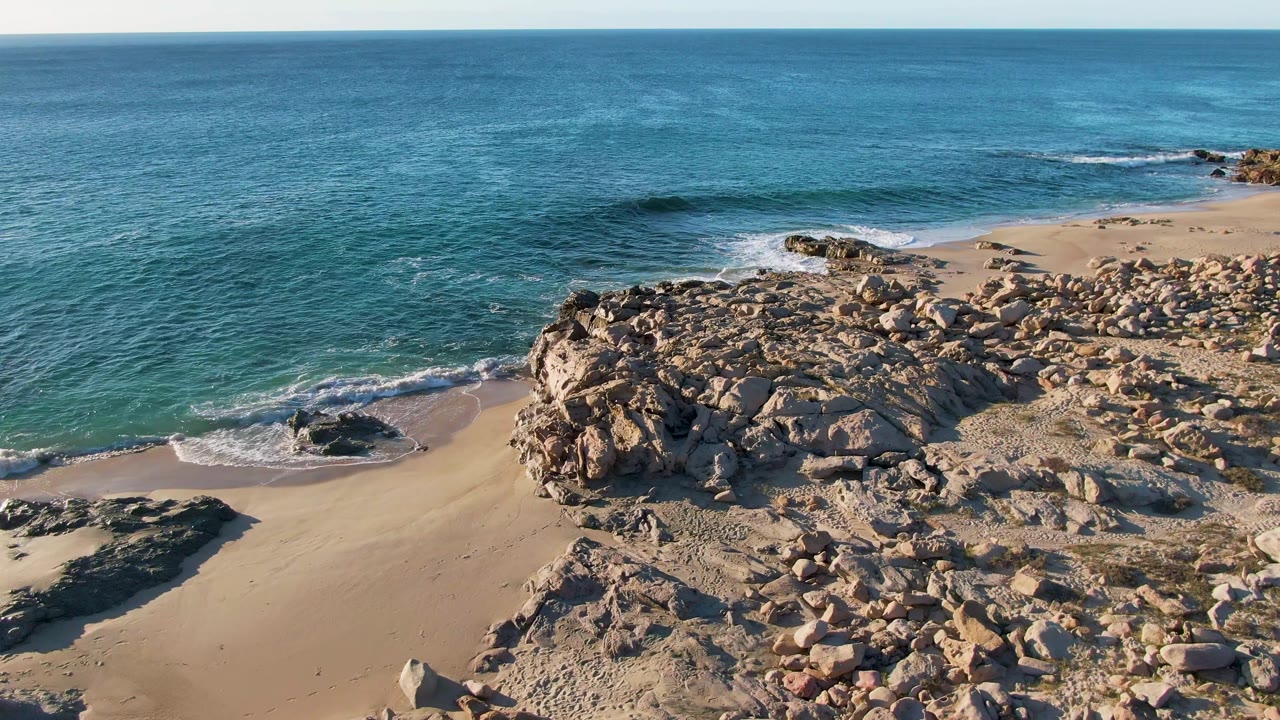 Wide aerial shoreline with rocks, sand, and calm ocean.