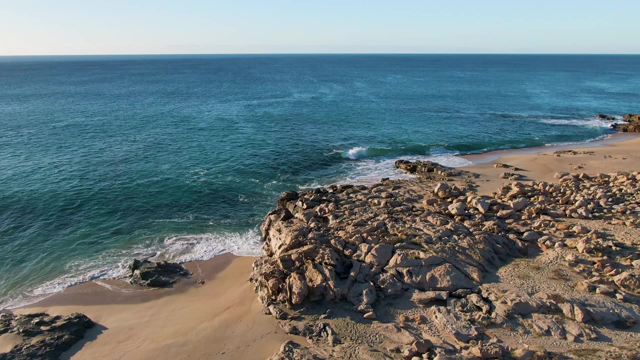 Aerial frame from Buzzards Beach East Cape BCS Mexico showing a broad rocky outcrop, tan sand, blue ocean, white foam at the shore, and no visible people.