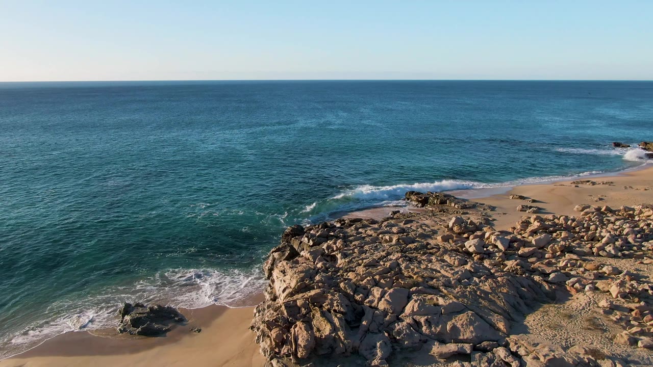 Buzzards Beach East Cape BCS Mexico appears from above with the rocky foreground pushed to the lower right, surf breaking along the sand, deep blue water beyond, and no visible people.