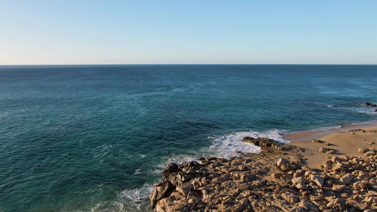 Mid-clip aerial frame over Buzzards Beach East Cape BCS Mexico showing darker teal water, a breaking wave near the rocks, sandy beach at right, and no visible people.