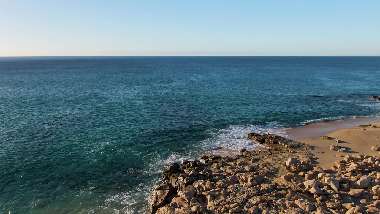 Aerial frame from Buzzards Beach East Cape BCS Mexico showing blue ocean, tan rocks, a small sandy beach, light surf, a clear horizon, and no visible people.