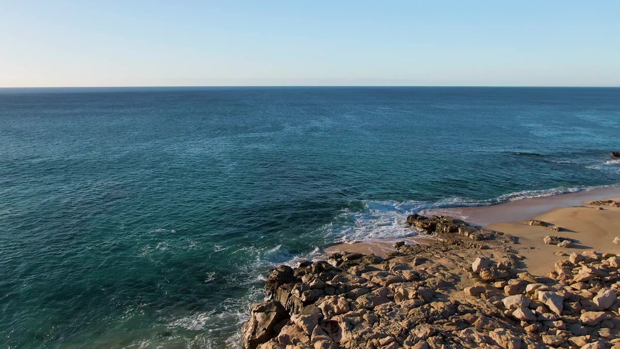 Aerial frame from Buzzards Beach East Cape BCS Mexico with blue-green water wrapping around foreground rocks, white foam on the beach edge, open sea, and no visible people.