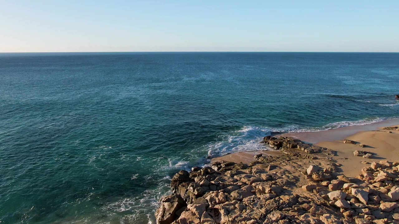 Aerial frame from Buzzards Beach East Cape BCS Mexico showing a rocky point in the lower frame, a curving sandy beach at right, textured blue water, and no visible people.