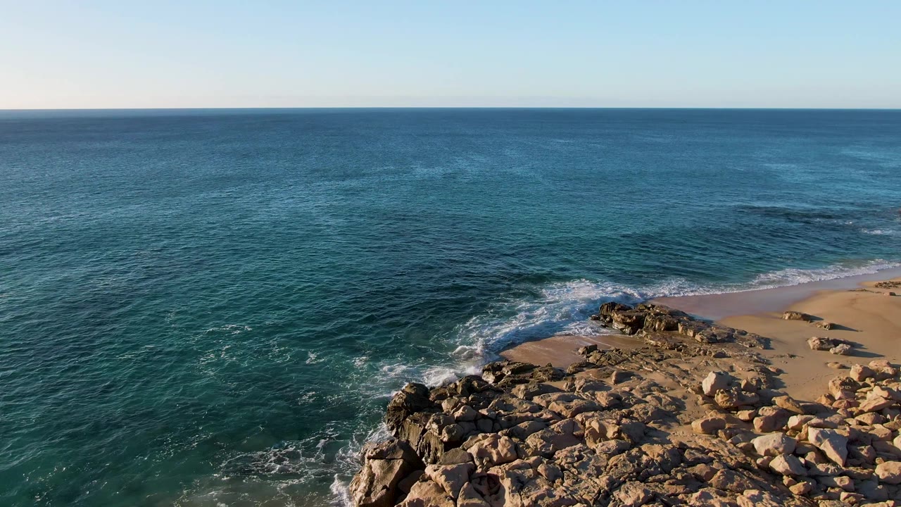 Wide aerial coastal view with ocean, rocks, surf, and beach.