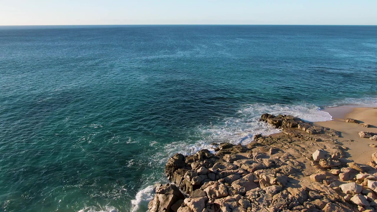 At 0:01, the Buzzards Beach aerial frame shows foam curling farther around the rocky point with turquoise water, sand to the right, and no visible people.
