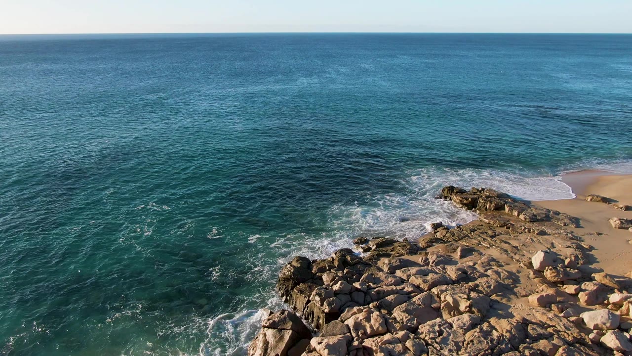 At 0:02, the aerial view over Buzzards Beach shows darker blue open water above, textured rocks below, a small sandy beach at right, and no visible people.