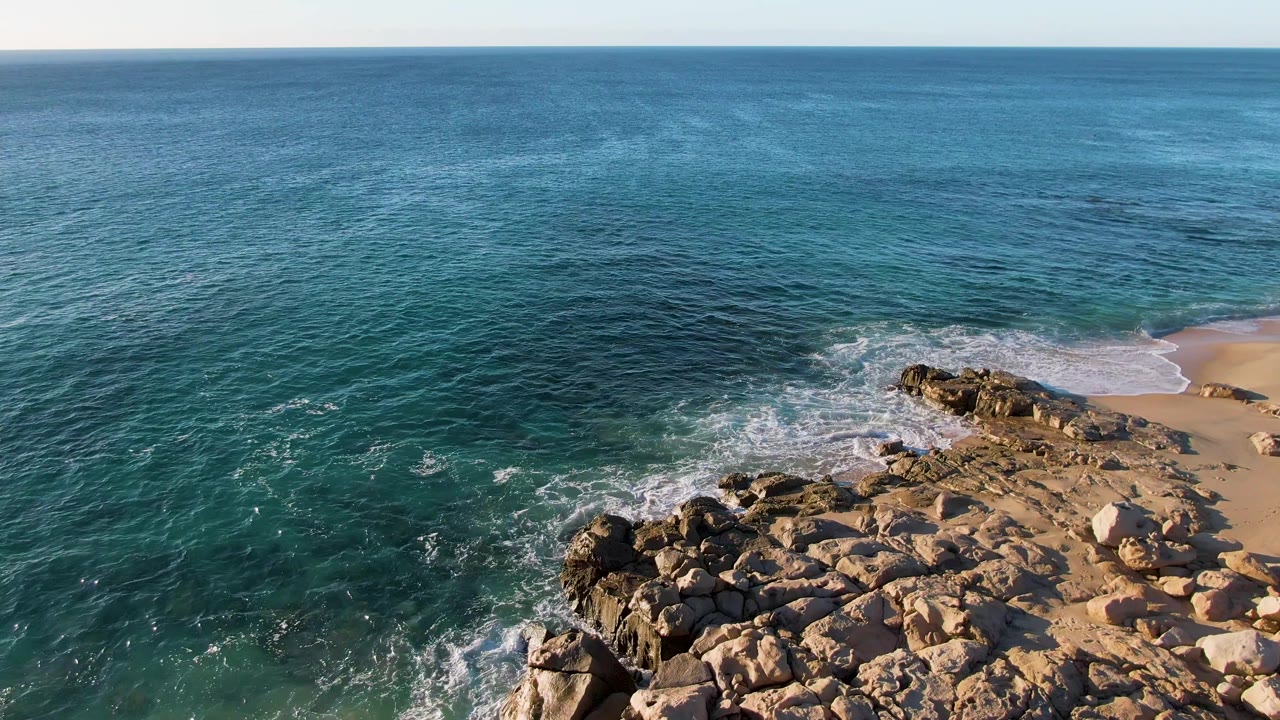 Blue-green ocean and white shorebreak beside tan rocks and beach sand.
