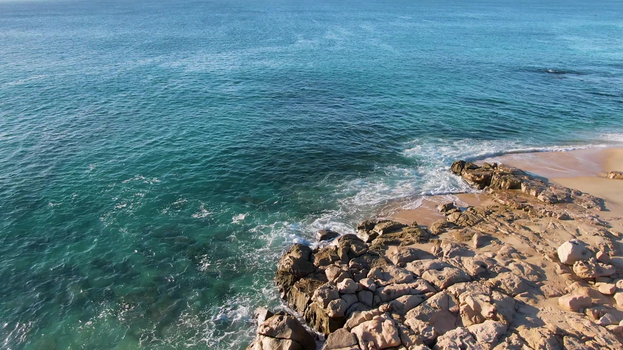 Aerial view of turquoise ocean washing over tan rocks beside a small sandy beach.