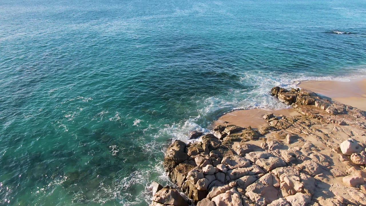 At Buzzards Beach East Cape BCS Mexico, a wave breaks against the upper-right rocks while the foreground rocks stay dry and sunlit, with no visible people.
