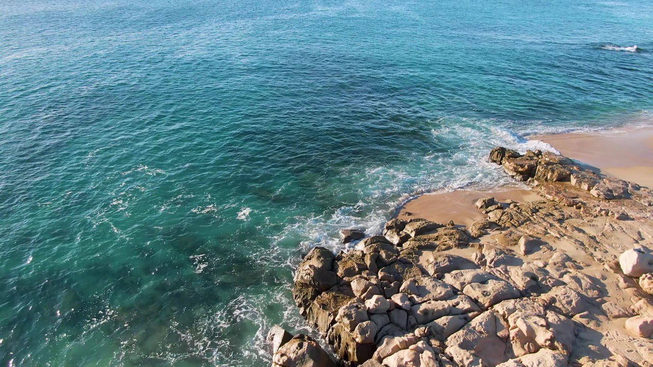 The mid-clip frame shows Buzzards Beach East Cape BCS Mexico from above, with more open turquoise water at left, a foamy shore break, and no visible people.