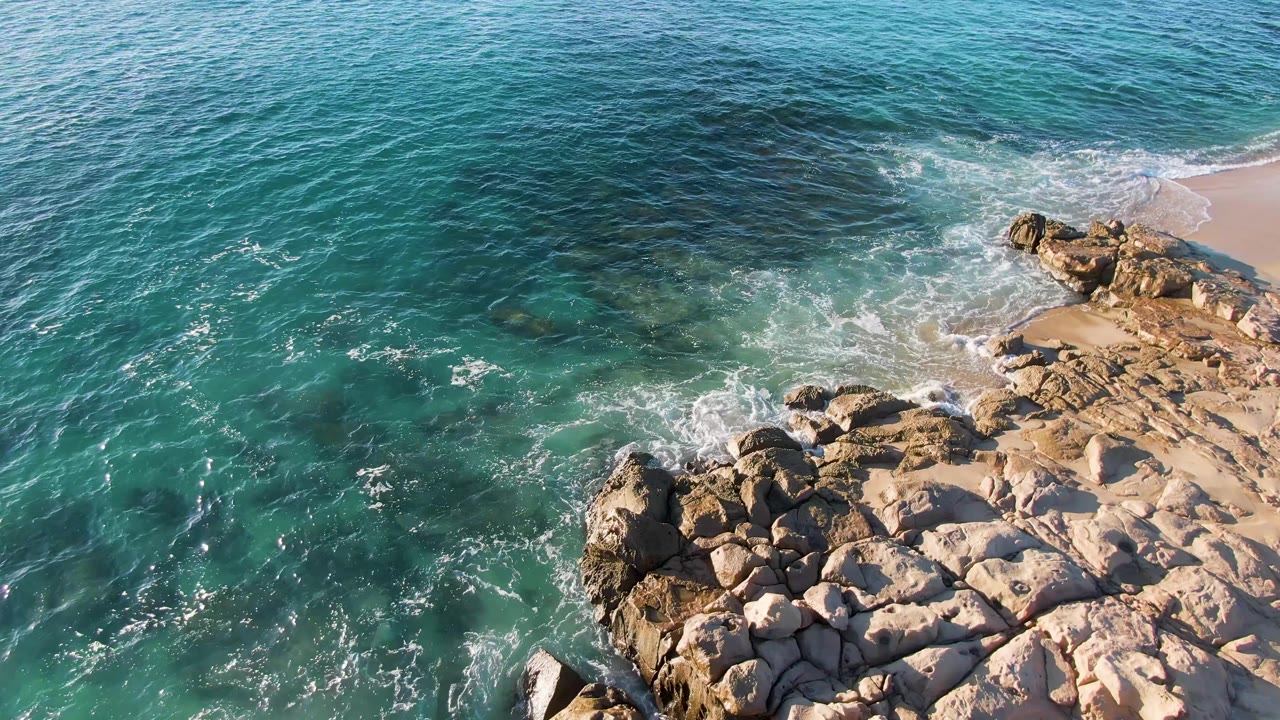 Aerial frame at Buzzards Beach East Cape BCS Mexico with blue-green water over visible seabed, foamy surf, shoreline rocks, and no visible people.