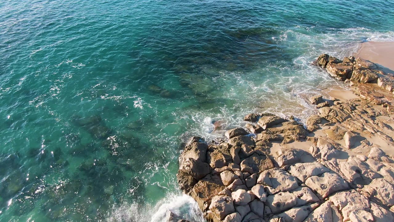 Aerial shoreline view at Buzzards Beach East Cape BCS Mexico where more open turquoise water fills the left side and rocks and sand sit along the right, with no visible people.