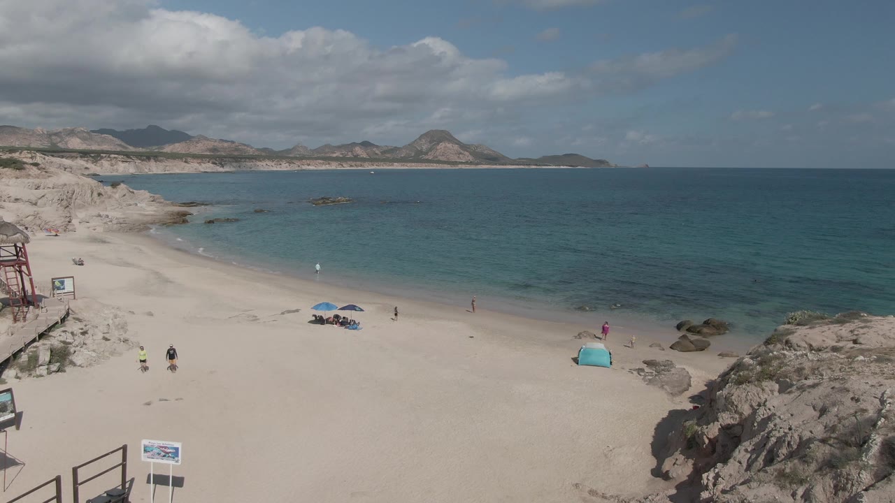 Aerial view of Playa El Arbolito Cabo Pulmo BCS Mexico with turquoise water, sandy beach, blue umbrellas, a turquoise tent, rocks, distant hills, and visible people near the shore.