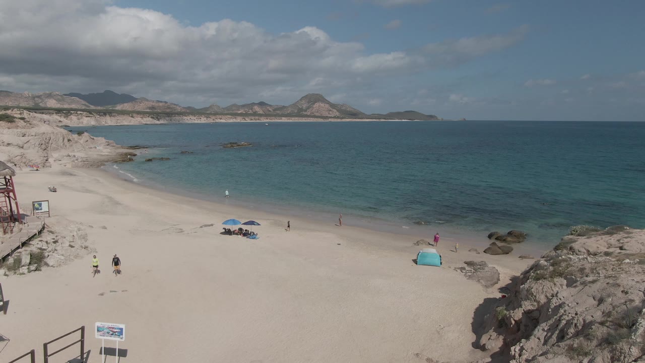 Wide aerial frame at Playa El Arbolito Cabo Pulmo BCS Mexico showing beachgoers walking on pale sand, blue umbrellas near the center, a turquoise tent by rocks, and calm turquoise water beyond.