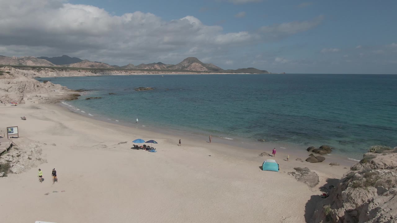 Aerial view of Playa El Arbolito Cabo Pulmo BCS Mexico with the shoreline sweeping across the frame, beachgoers spaced along the sand, blue umbrellas, a small turquoise tent, rocks, and hills under clouds.