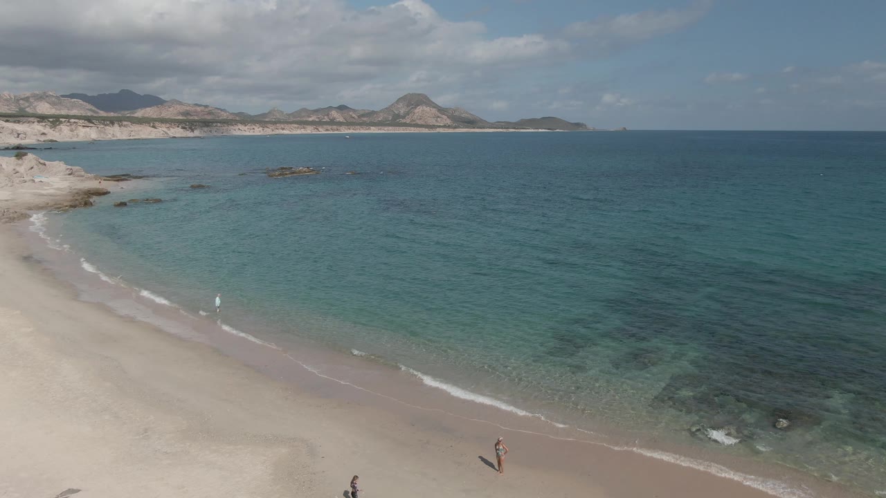 Aerial shoreline view with clear water, small people, and mountains in the distance.