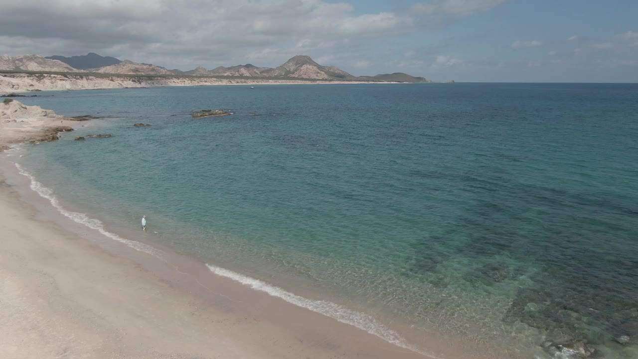 Wide aerial frame of Playa El Arbolito with sandy beach along the lower left, one small person near the wash, rippled turquoise water, rocky shallows, and mountains on the horizon.