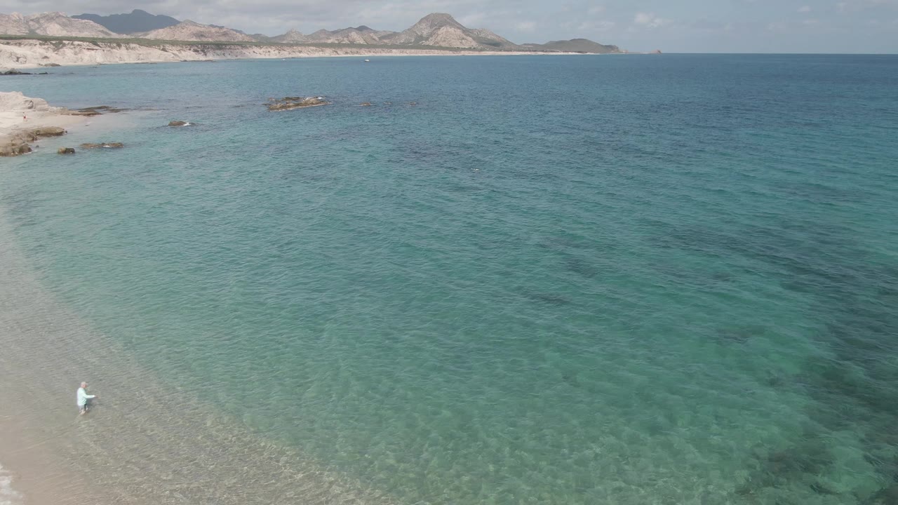 Aerial view at Playa El Arbolito, Cabo Pulmo, Mexico, with a person in white near the lower left shoreline, shallow turquoise water, small rocks, and distant hills.