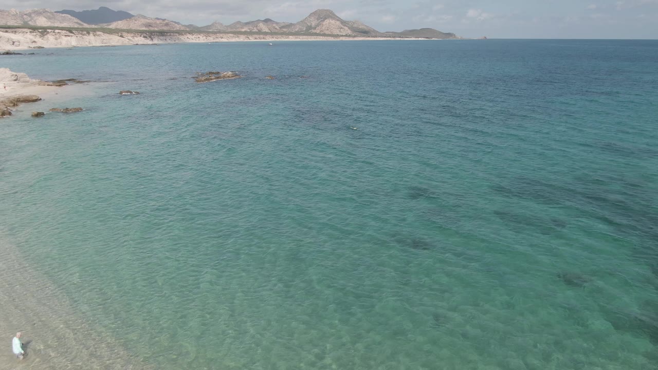 Turquoise coastal water with beach, rocks, and distant hills.