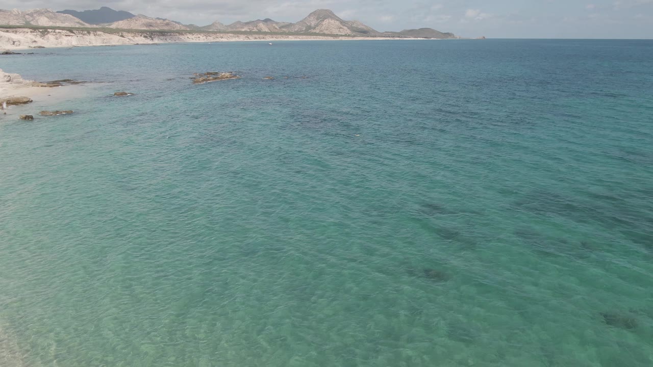 Aerial view at Playa El Arbolito, Cabo Pulmo, Mexico, with no visible people, clear turquoise shallows in the foreground, a pale beach at left, rock patches, and distant hills.