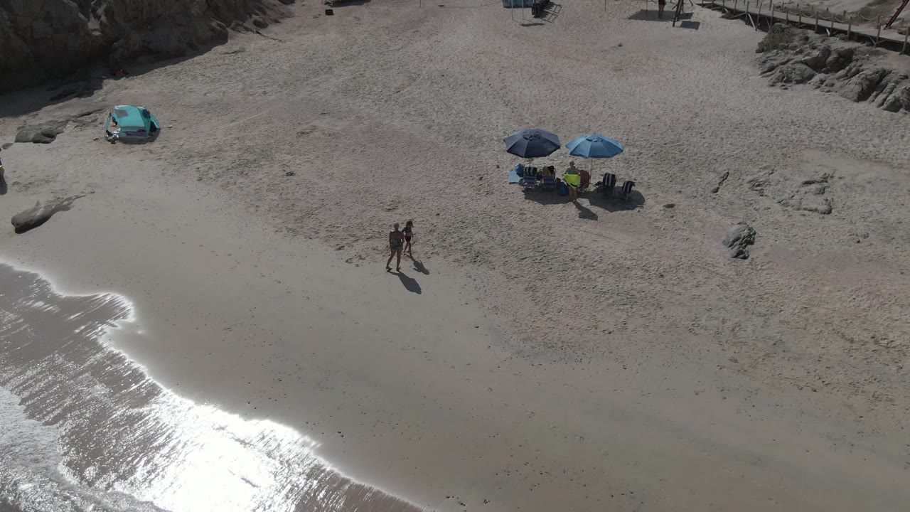 At Playa El Arbolito Cabo Pulmo BCS Mexico, two people walk on pale sand above a bright reflective wave edge, with a turquoise car and blue umbrellas higher on the beach.