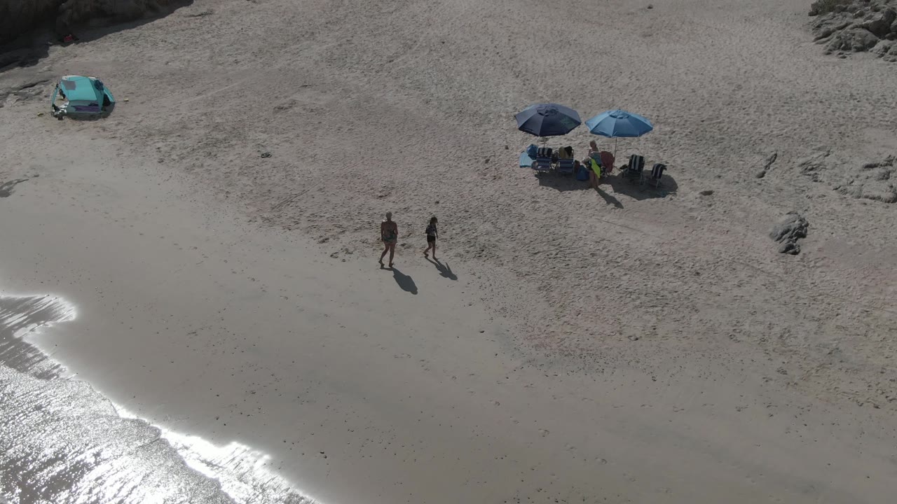 At Playa El Arbolito Cabo Pulmo BCS Mexico, two people continue across the sand beside a curved strip of bright surf glare, with umbrellas and seated beachgoers farther up the beach.