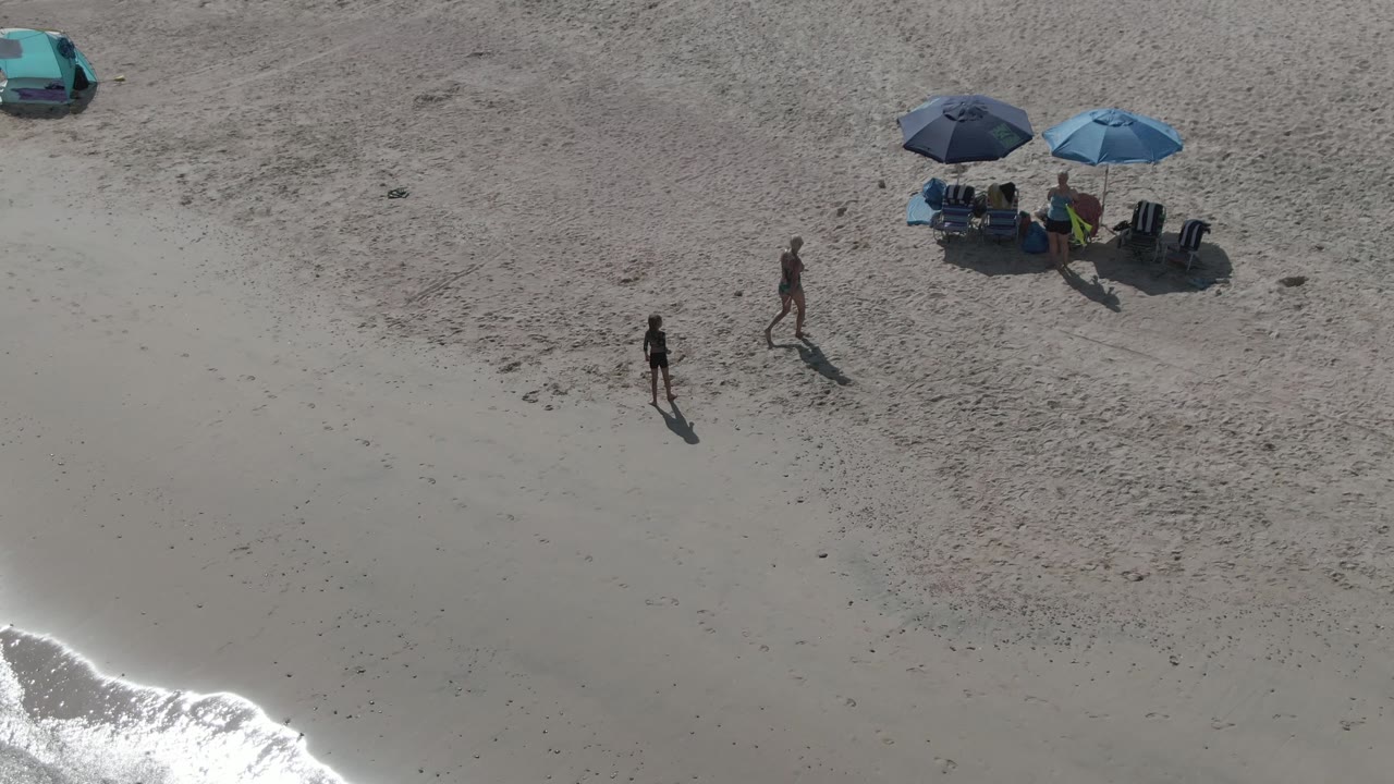 Overhead frame at Playa El Arbolito shows an adult and child walking on pale sand below blue umbrellas and a teal car, with bright surf entering the lower left.