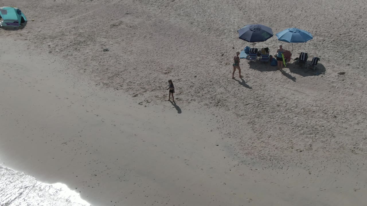At 0:02, the child remains lower on the sand while the adult is closer to the blue umbrellas and chairs at Playa El Arbolito, with a teal car near the top edge.