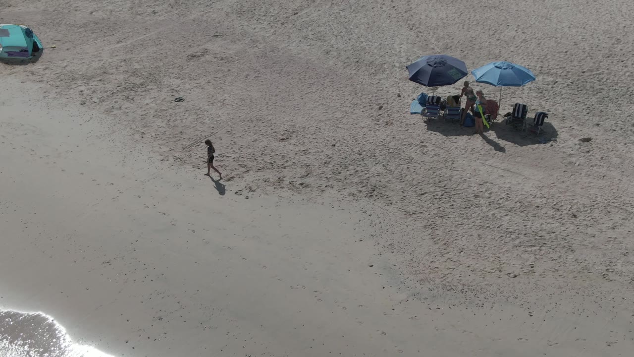 Overhead view of Playa El Arbolito in Cabo Pulmo, Mexico, with two umbrellas over beach chairs, a teal car on the sand, a walker near the shore, and surf glinting at the lower left.