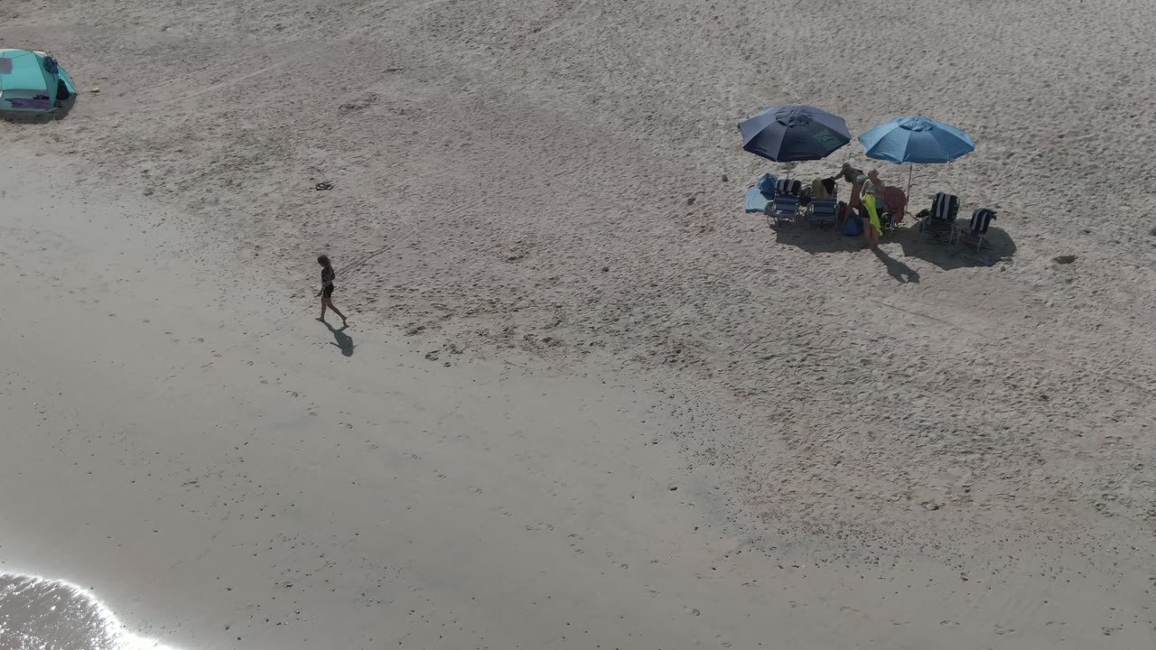 Aerial beach view with a walker crossing pale sand near umbrellas and a teal car.
