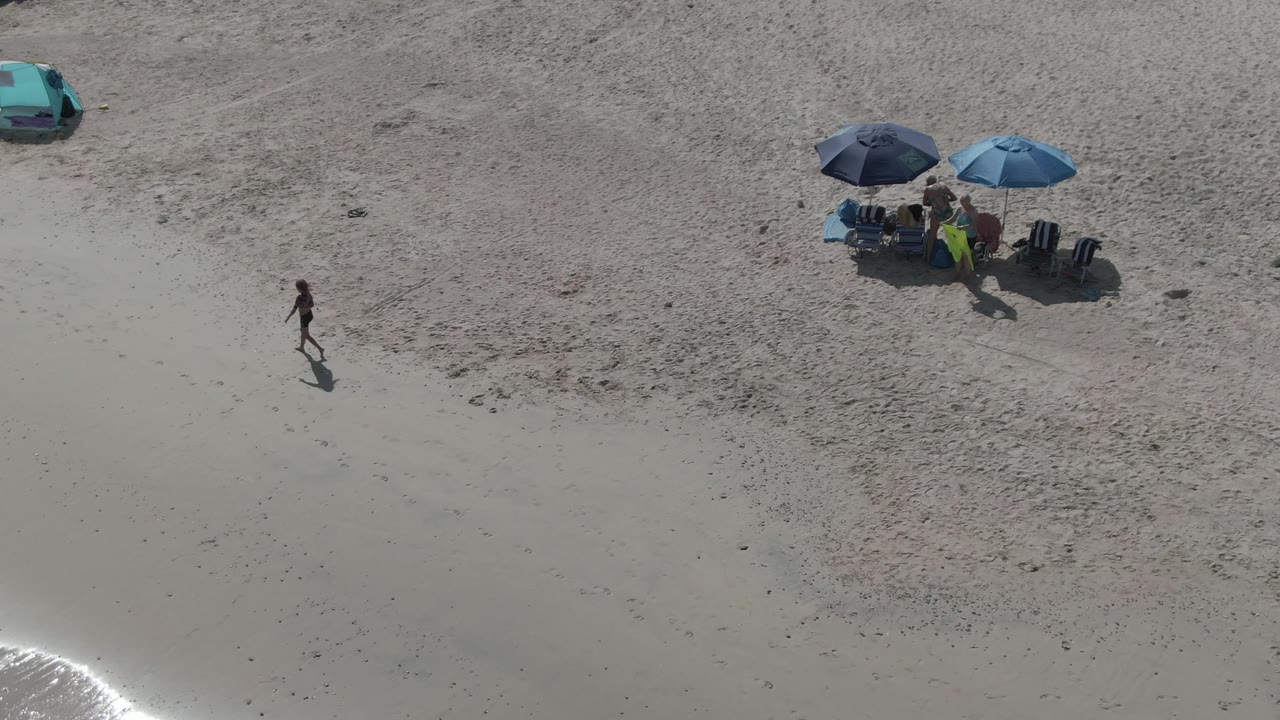 Top-down view of Playa El Arbolito in Cabo Pulmo, Mexico, with the walker centered lower in the frame, two umbrellas and seated beachgoers near the upper right, and a teal car on the sand.