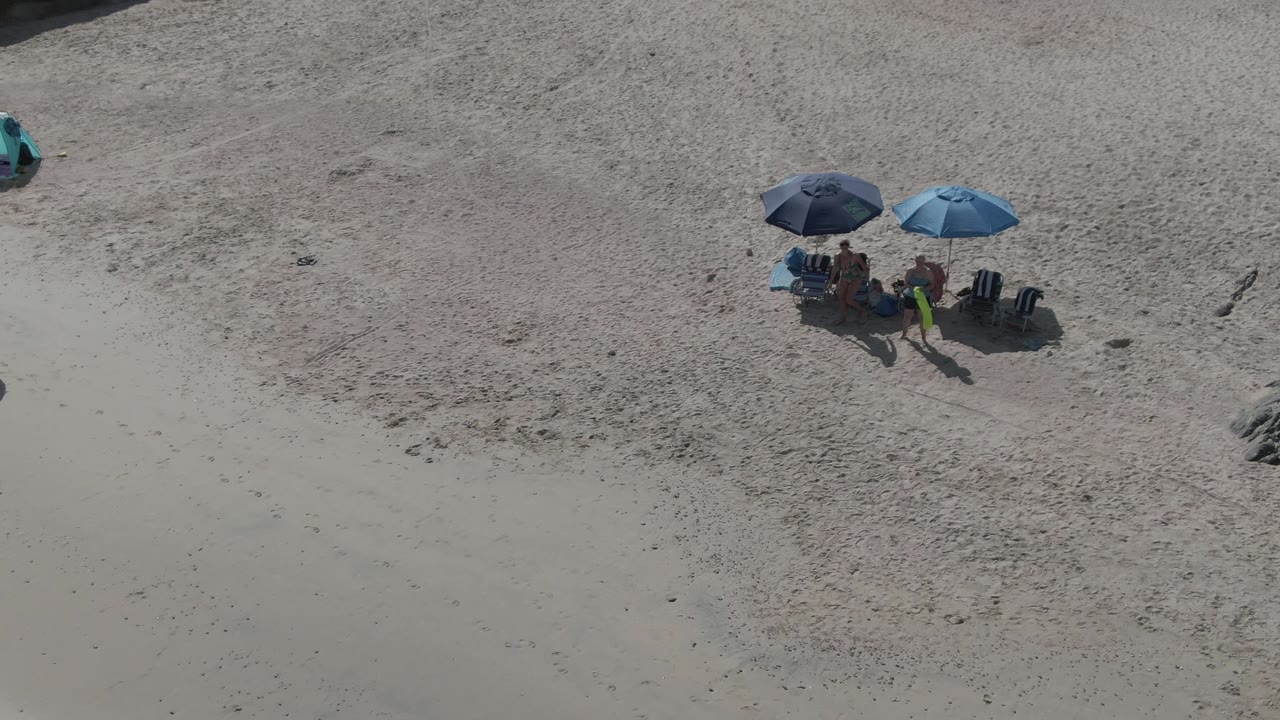 People standing and sitting around two beach umbrellas on sand.