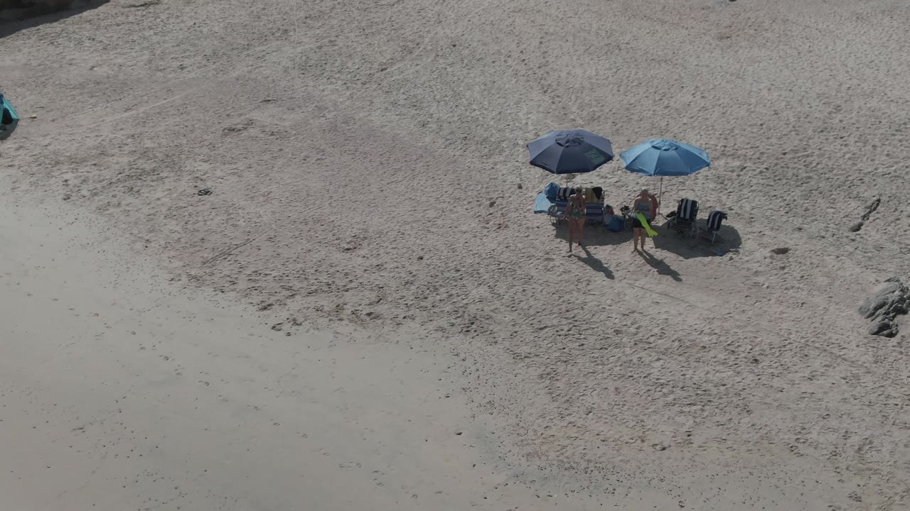 Overhead Playa El Arbolito Cabo Pulmo view with two blue umbrellas, several beachgoers around chairs, long shadows, and textured sand extending across the background.