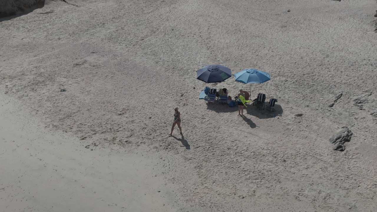 Aerial frame from Playa El Arbolito Cabo Pulmo, Mexico showing a person walking across pale sand below a group seated under two blue umbrellas with rocks nearby.
