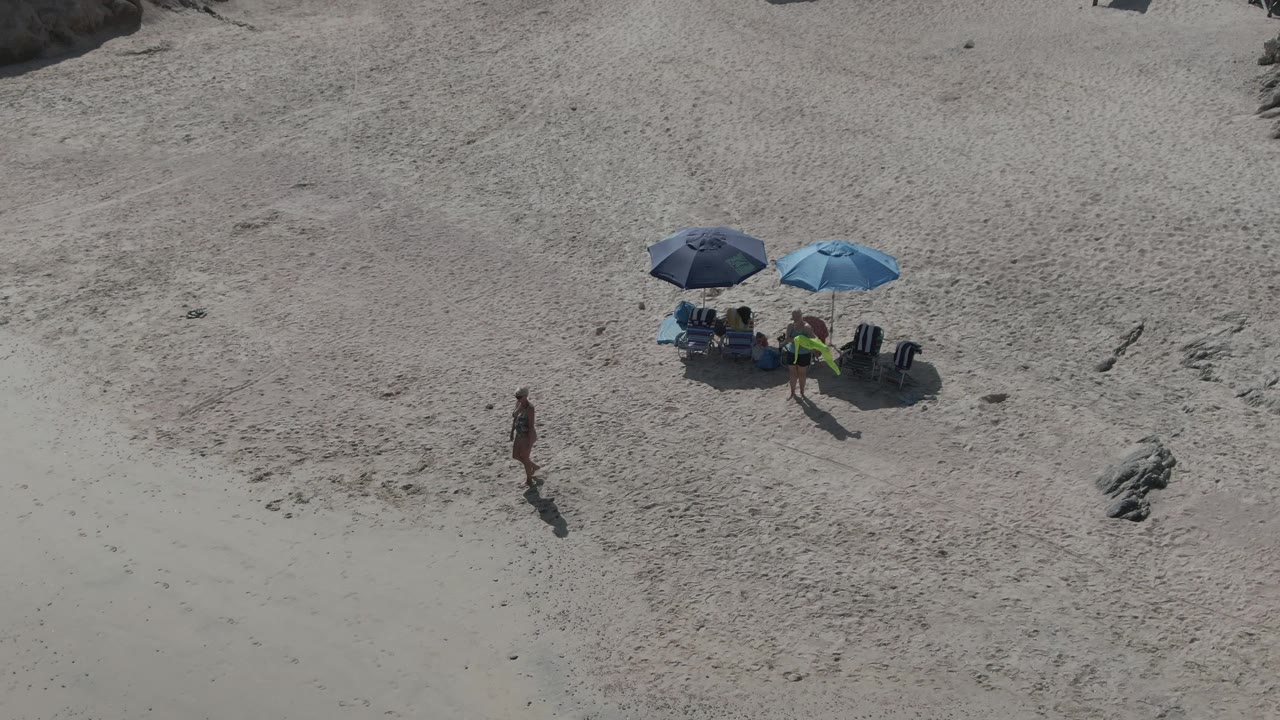 Person in swimwear walks on sandy beach near two blue umbrellas.