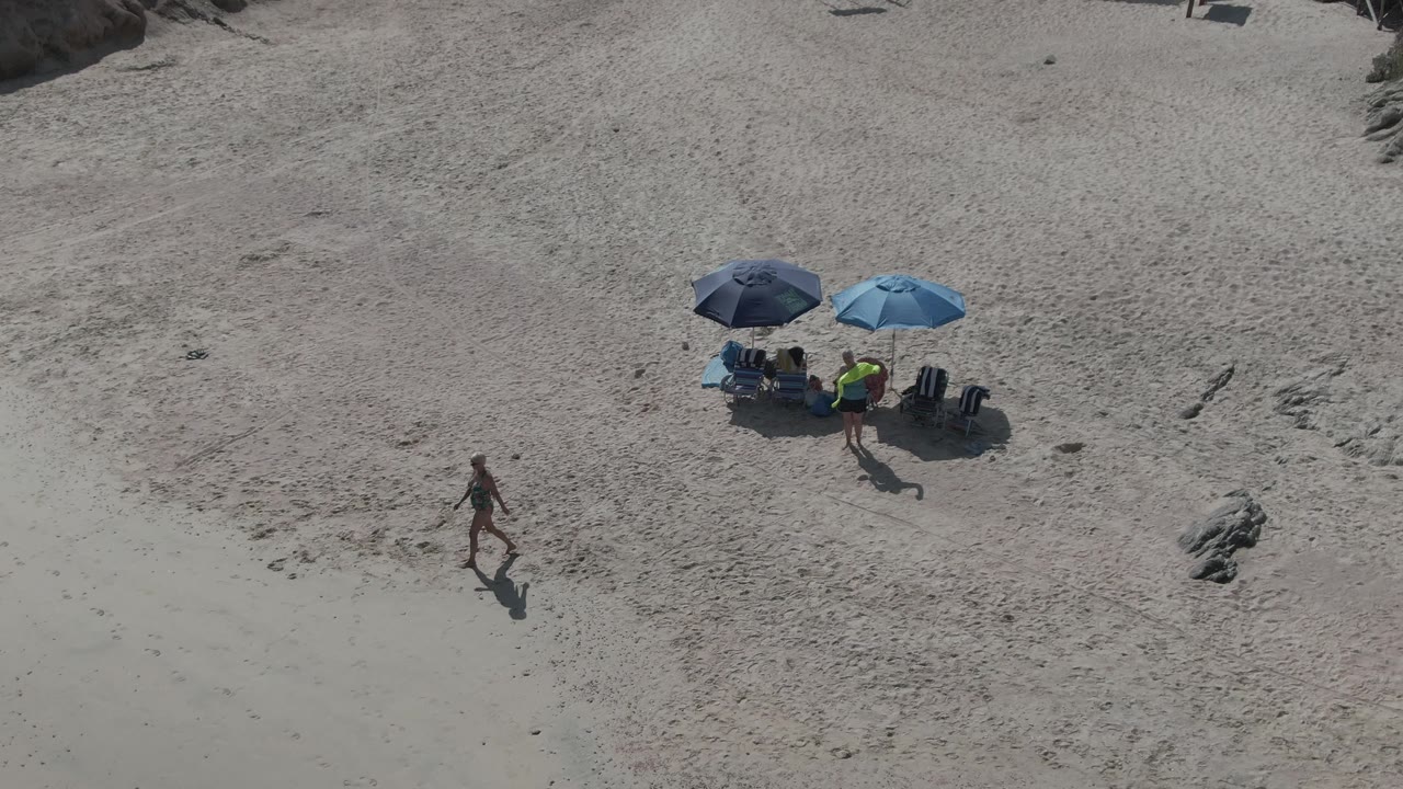 Overhead Playa El Arbolito Cabo Pulmo, Mexico beach frame with the walking person left of the umbrella group, empty sand around them, and a small rock cluster nearby.