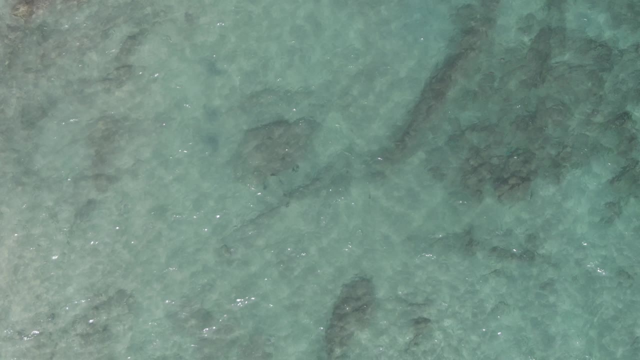 Aerial-looking view of turquoise water at Playa El Arbolito Cabo Pulmo BCS Mexico, with mottled seabed tones and rock shapes visible through the rippled surface, no visible people.