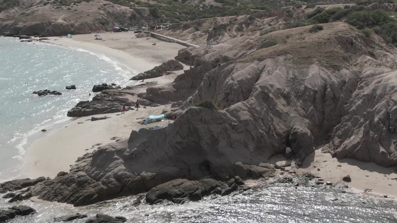 Aerial frame at Playa El Arbolito showing a sandy cove below gray cliffs, turquoise water on the left, small waves at the shore, and tiny people on the lower beach.