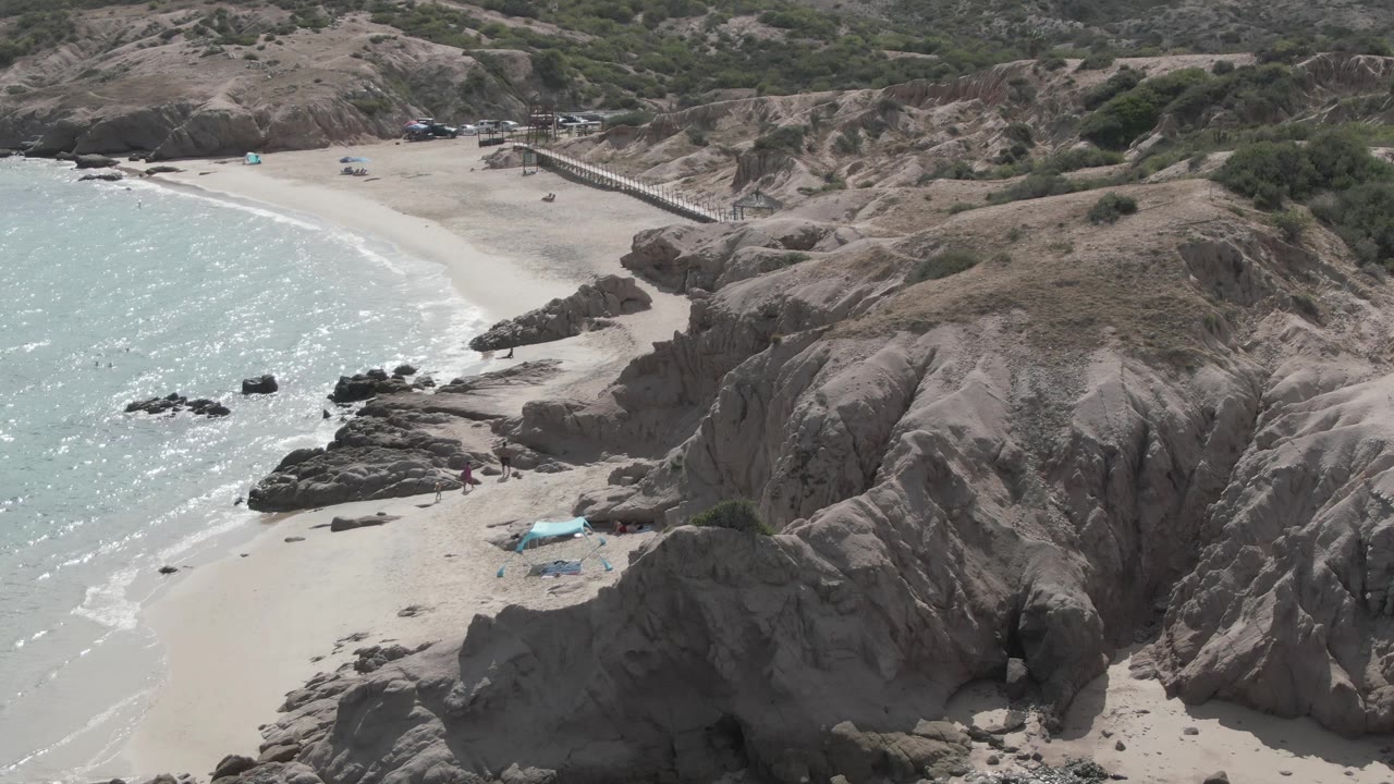 High aerial frame at Playa El Arbolito showing more of the cliff face on the right, sparkling turquoise water on the left, pale sand below, and small people near the rocks.