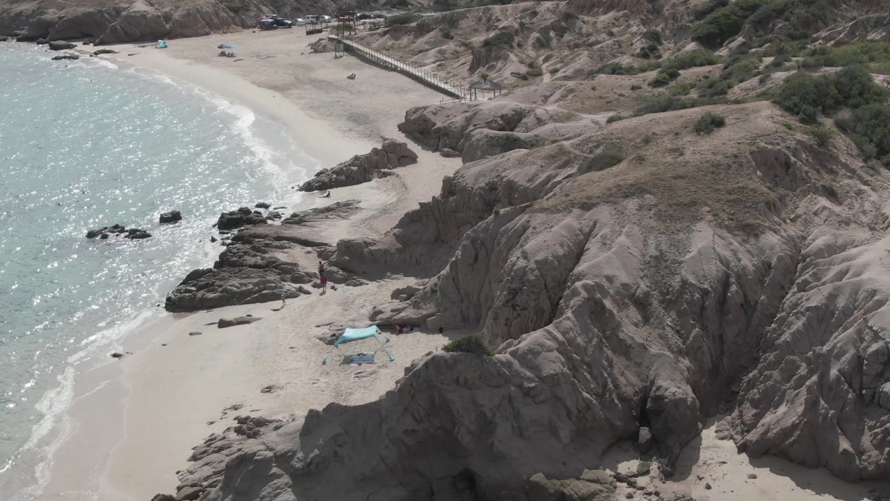 Aerial frame over Playa El Arbolito shows tan rock outcrops beside pale sand, turquoise water, a blue shade tent, and a few small people on the beach.