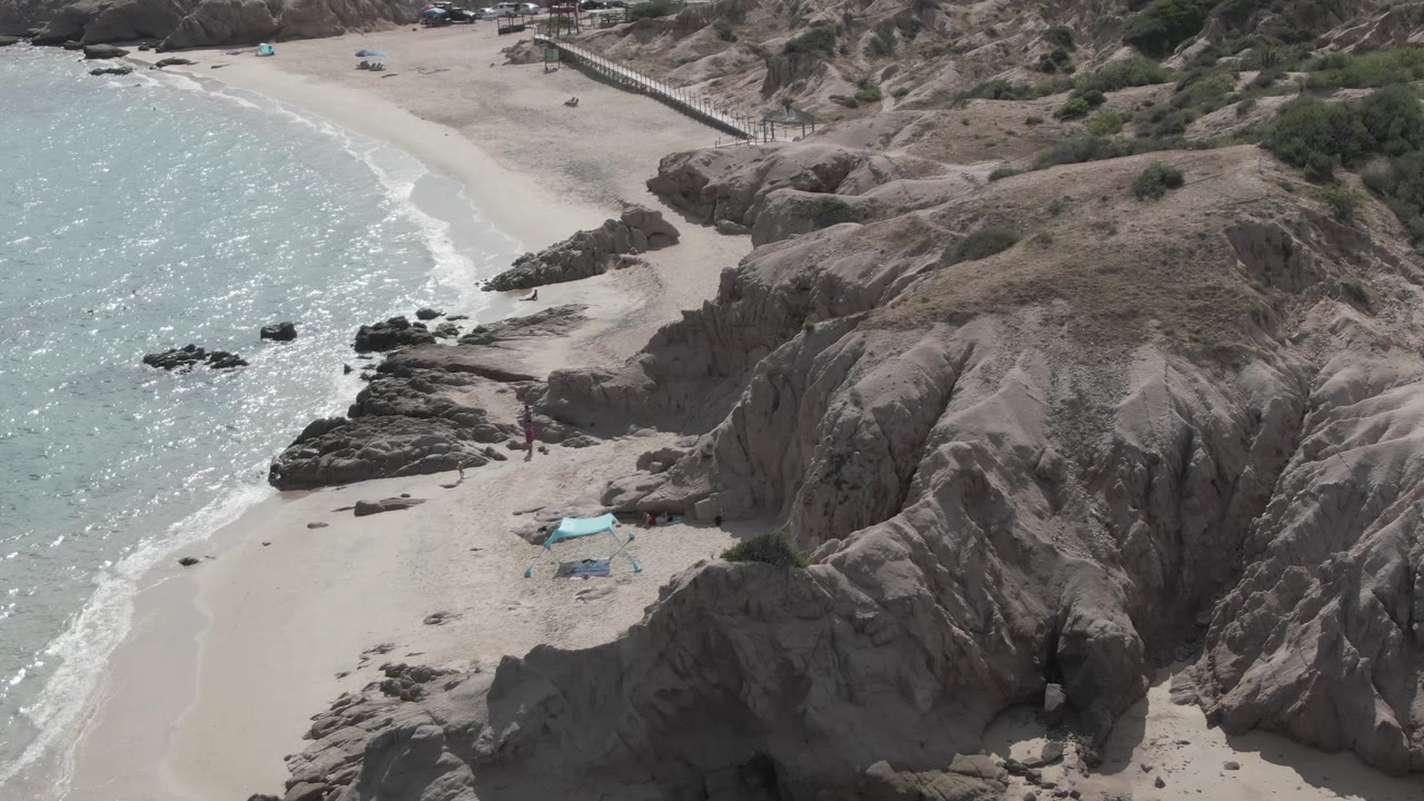 Drone view of sandy cove with cliffs, waves, and beachgoers