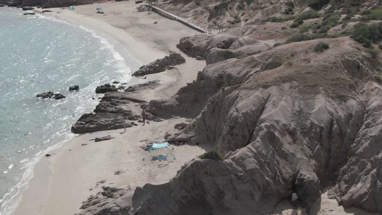 At 0:02, Playa El Arbolito appears from a slightly shifted overhead angle, with sunlit turquoise water, dark rocks in the surf, sand, a blue tent, and tiny people near the rocks.