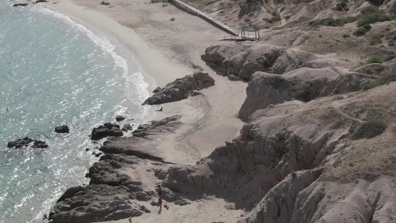 At 0:00, Playa El Arbolito appears from above with turquoise water at left, pale sand below, rounded rocks through the center, and a raised walkway near the upper beach.