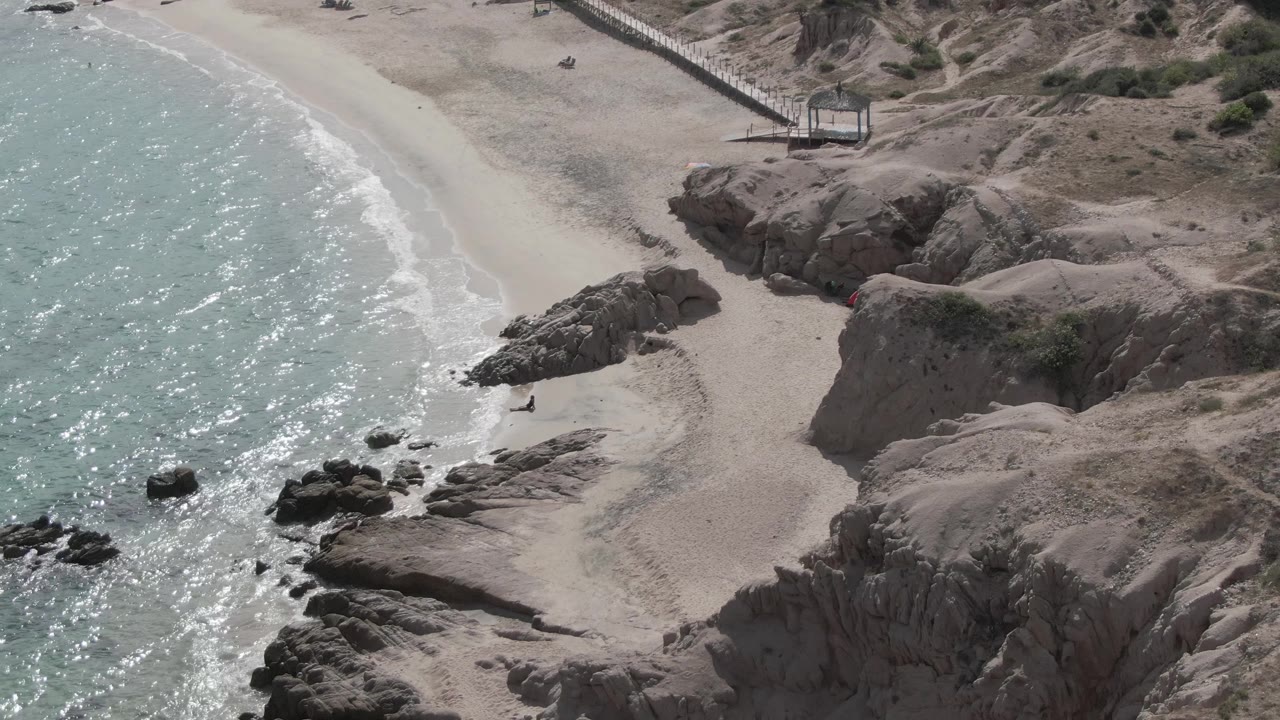 Overhead coastal view with rocky outcrops, sandy cove, small people, and breaking waves.